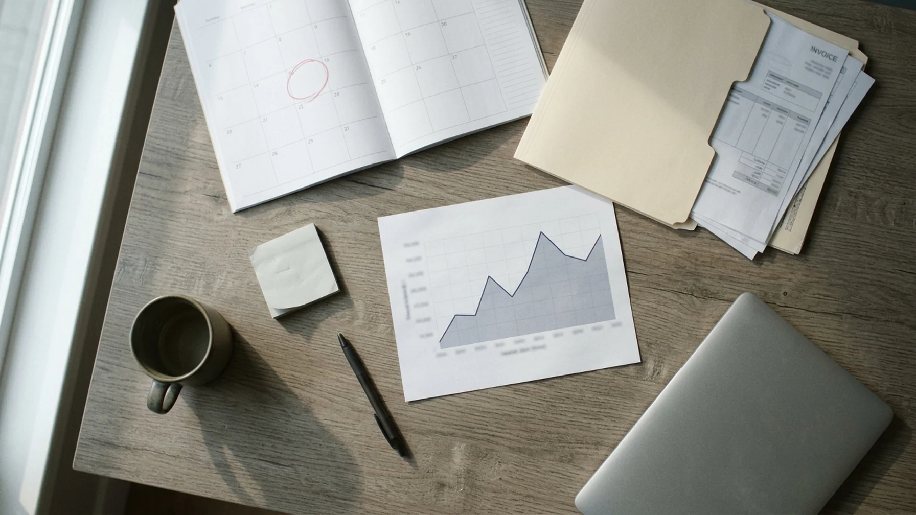 Finance manager marking a calendar three months ahead beside a folder of supplier invoices, with a currency chart printout on the desk